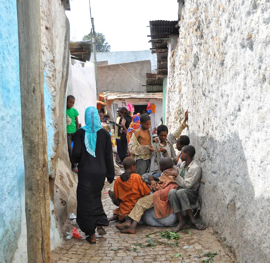 A narrow cobbled alley inside Harar's walled old town, lined with plain whitewashed and painted walls, with people sitting and walking through it.