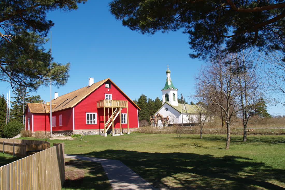The red wooden Kihnu museum building with the white church and its onion dome behind it.